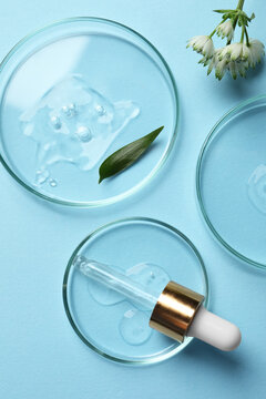 Petri dishes with samples of cosmetic oil, pipette and flowers on light blue background, flat lay