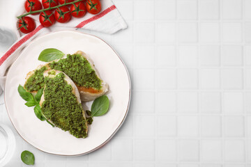Delicious chicken breasts with pesto sauce, basil and tomatoes on white tiled table, flat lay. Space for text