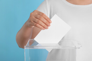 Woman putting her vote into ballot box on light blue background, closeup