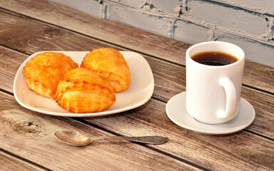 A cup of hot black coffee on a saucer, next to it is a spoon and a plate with three fresh curd buns on a wooden table.