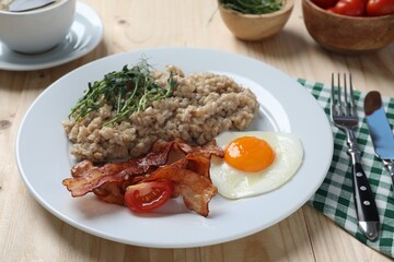 Delicious boiled oatmeal with fried egg, bacon and tomato served on wooden table, closeup