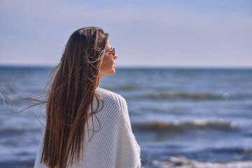 Obraz premium Portrait of attractive brunette girl with long hair posing on a deserted beach. She wears white shorts, a top, a long white cape. She is looking to the camera.
