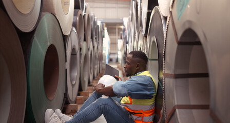 A young man works in a warehouse storing rolls of metal sheet material. Sit down break and wipe off sweat a little before continuing with work.
