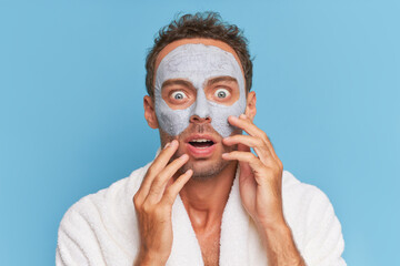 Close-up of young man face with clay cosmetic mask, fear expression on his face holding hands at his cheeks, spa treatments concept, copy space