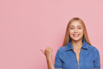 Pretty fair-haired woman standing on pink background dressed in blue denim dress pointing thumb...