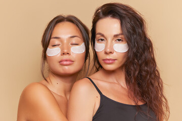 Two cute brunette girls make cosmetic procedures on their faces, each has eye patches on face,...