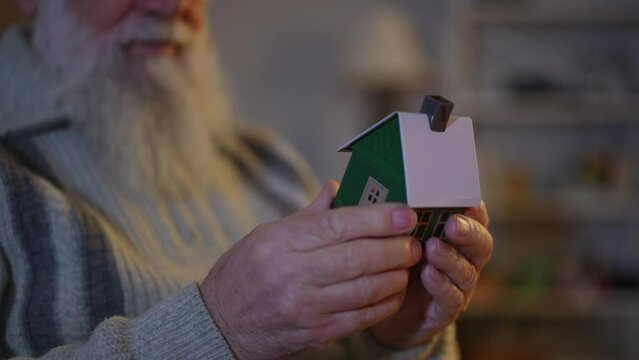 An elderly man with a beard carefully examines from all sides a toy house made of thin metal