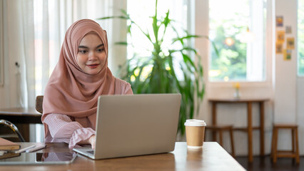 A beautiful Asian-Muslim woman is working on her laptop, working remotely at a coffee shop.