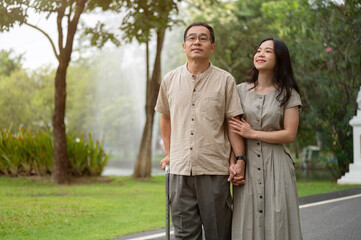 A smiling Asian daughter and her dad with a walking stick are walking in the park together.