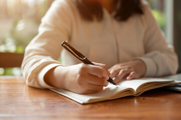 Cropped image of an Asian woman in casual clothes writing something in her book at a table indoors.