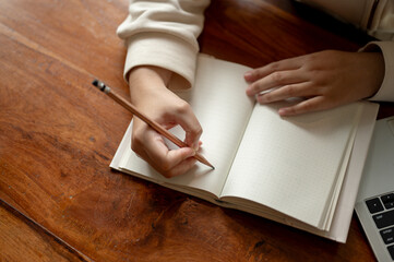 Top view of a woman holding a pencil, writing in a book at a wooden table. Blank empty pages mockup.
