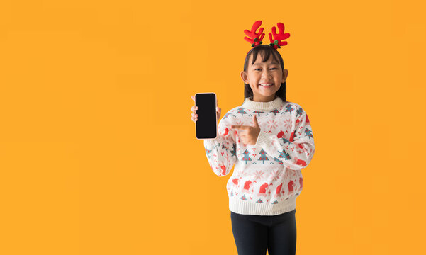 Cheerful Asian little girl wearing a Christmas sweater with reindeer horns, Happy smiling while holding smart phone front blank screen and hand pointing, isolated on yellow background