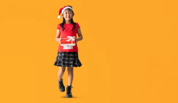 Happy smiling Asian child girl in santa claus hat Hands holding xmas gift box happy new year, Full body isolated on yellow background