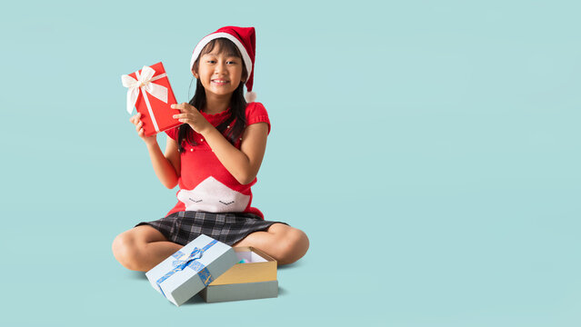 Portrait of cheerful happy Asian little girl woman in santa claus clothes with gift box, Sitting Hands holding xmas gift box happy new year, isolated on pastel plain light blue background