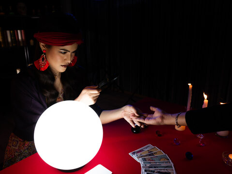 Asian astrologer woman holding customer hand and use palmistry skill with magnifying glass for zoom the detail before predict destiny and future life in the dark room