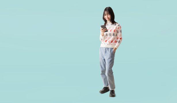 Happy smiling asian teenager girl standing using smart phone, Wearing a Christmas sweater, Portrait full body isolated on pastel plain light blue background