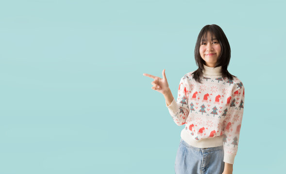 Happy smiling asian teenager girl pointing at something wearing a Christmas sweater, isolated on pastel plain light blue background
