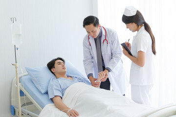 Senior doctor and young male patient who lie on the bed while checking pulse, consult and explain with nurse taking note and supporting in hospital wards. - obrazy, fototapety, plakaty