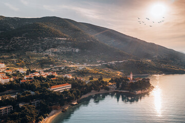 Picturesque aerial view of Brac, Croatia during golden hour. The setting sun casts a warm glow over the coastal village nestled between lush green hills and the serene Adriatic Sea. 