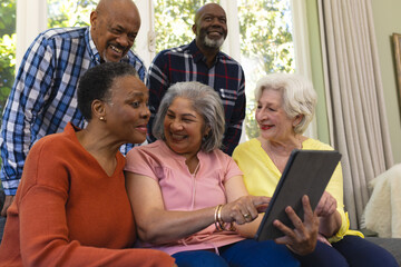 Happy diverse group of senior friends using tablet and laughing in sunny living room