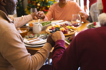 Hands of diverse group of senior friends saying grace at christmas dinner in sunny dining room