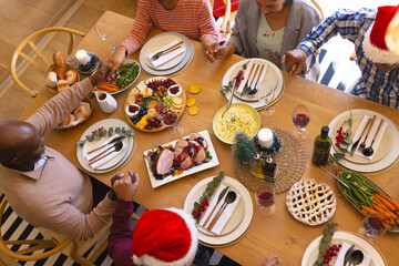 Diverse group of senior friends saying grace at christmas dinner in sunny dining room at home