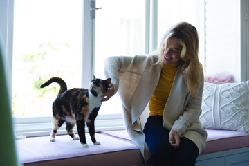Happy caucasian woman sitting on windowsill and stroking cat in sunny room at home