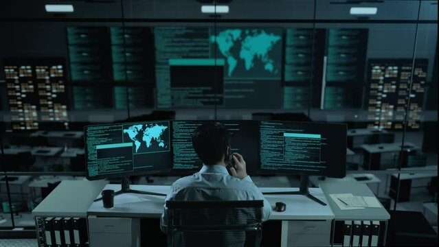 Back View Of Asian Man Developer Talking On Mobile Phone While Write Code With Multiple Computer Screens In The Office
