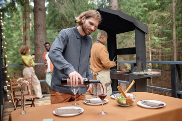 Young man pouring wine in glasses during picnic while his friends cooking meat on grill in background