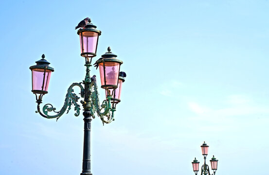 Lantern on the street of Venice. The famous pink lights of Venice. Four lamps with pink glass are on an ornate black metal lamp post. Blue cloudless sky. Nice background, postcard