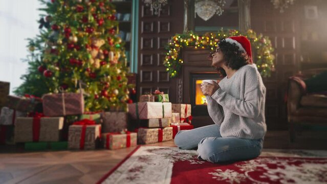 Relaxing african american woman in Santa hat drinking beverage. Girl sitting near Christmas tree fireplace smiling, feeling harmony New Year spirit at home in living room. Celebrating winter holidays.