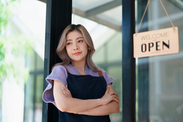 open a small business Portrait of happy Asian woman wearing apron holding welcome we are open sign at front of cafe.