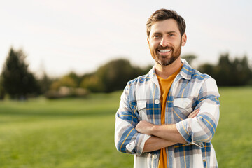 Portrait of smiling handsome middle aged man farmer wearing s stylish t shirt holding arms crossed looking at camera standing in green field, copy space. Successful business concept - obrazy, fototapety, plakaty