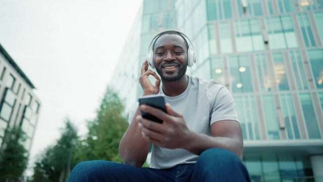 African american man music lover in headphones enjoying listening music on smartphone sitting on city street outdoors. Positive smiling guy melomaniac rhythmically nodding head relaxing moving body.