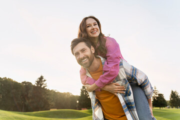 Portrait of attractive smiling middle aged couple looking at camera in green park. Happy man and woman carry on shoulders spending time together on nature.  Date, love, relationship, travel concept 
