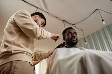 Low angle view of young barber using trimmer to do stylish haircut for customer