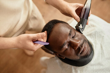 African American customer sitting with his eyes closed while barber shaving his beard using shaver and comb