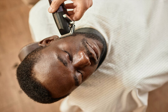 Top view of African American young man lying in chair with closed eyes while barber shaving his beard with shaver
