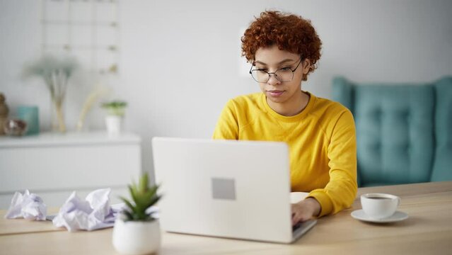 African american girl student writing educational project on paper sheets crumpling bad ones sitting at table on workplace at home. Curly creative woman working on documents. Education, study, learn.