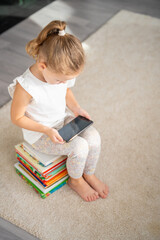 Little girl sits on a stack of children's fairy-tale books and watches cartoons on her smartphone