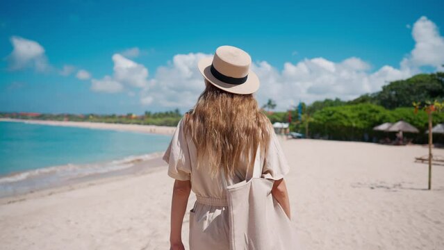 Romantic blonde woman tourist in straw hat walking to ocean sandy beach in sunny day, rear view. Female lady enjoying nature on vacations summer holidays. Travel, tourism, journey, rest relax concept.
