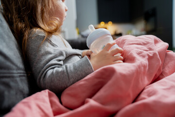 Small toddler girl with long hair is sick staying home covered in pink blanket while holding small...