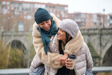 Happy dominican girl giving a piggyback ride to her girlfriend with a coffee cup at street in...