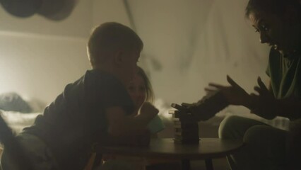 Little boy playing with mother and sister accidentally drops entire tower onto table. Mom and children surprised with end of game and cover mouths. Family board game night