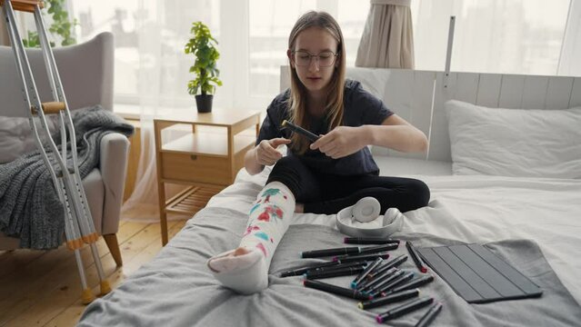 Teenage girl after injury with broken leg in orthopedic cast painting pictures felt tips sitting in bed at home. Teen in glasses drawing on cast. Recovery of damaged leg, forced sick leave concept.