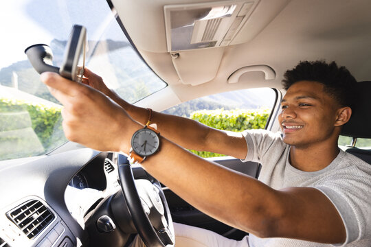 Happy african american man adjusting rearview mirror in car on sunny day