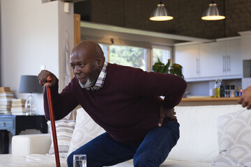 Pained african american senior man with walking stick standing up from couch at home holding back