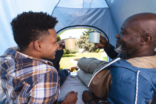 Happy african american father and adult son in tent drinking a toast with cups of coffee