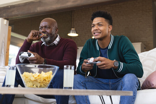Happy african american father and adult son at home playing video game with gamepads