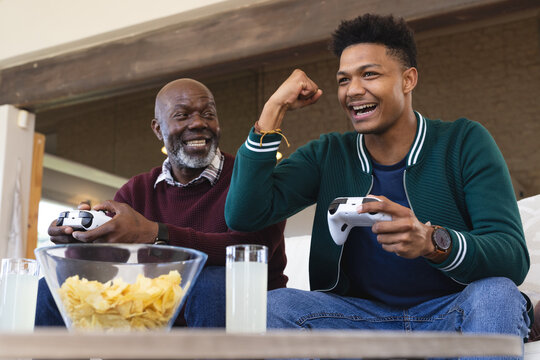 Happy african american father and adult son playing video game with gamepads and celebrating victory